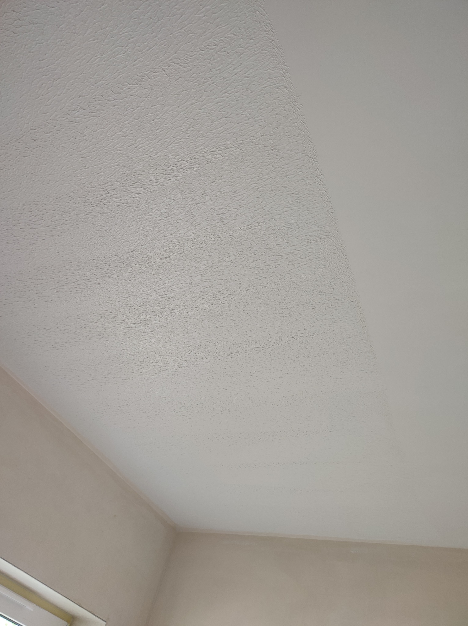 Częściowo pomalowany sufit z widoczną fakturą, kontrastujący z gładką, świeżo pomalowaną powierzchnią, widoczne łączenie ścian i sufitu.