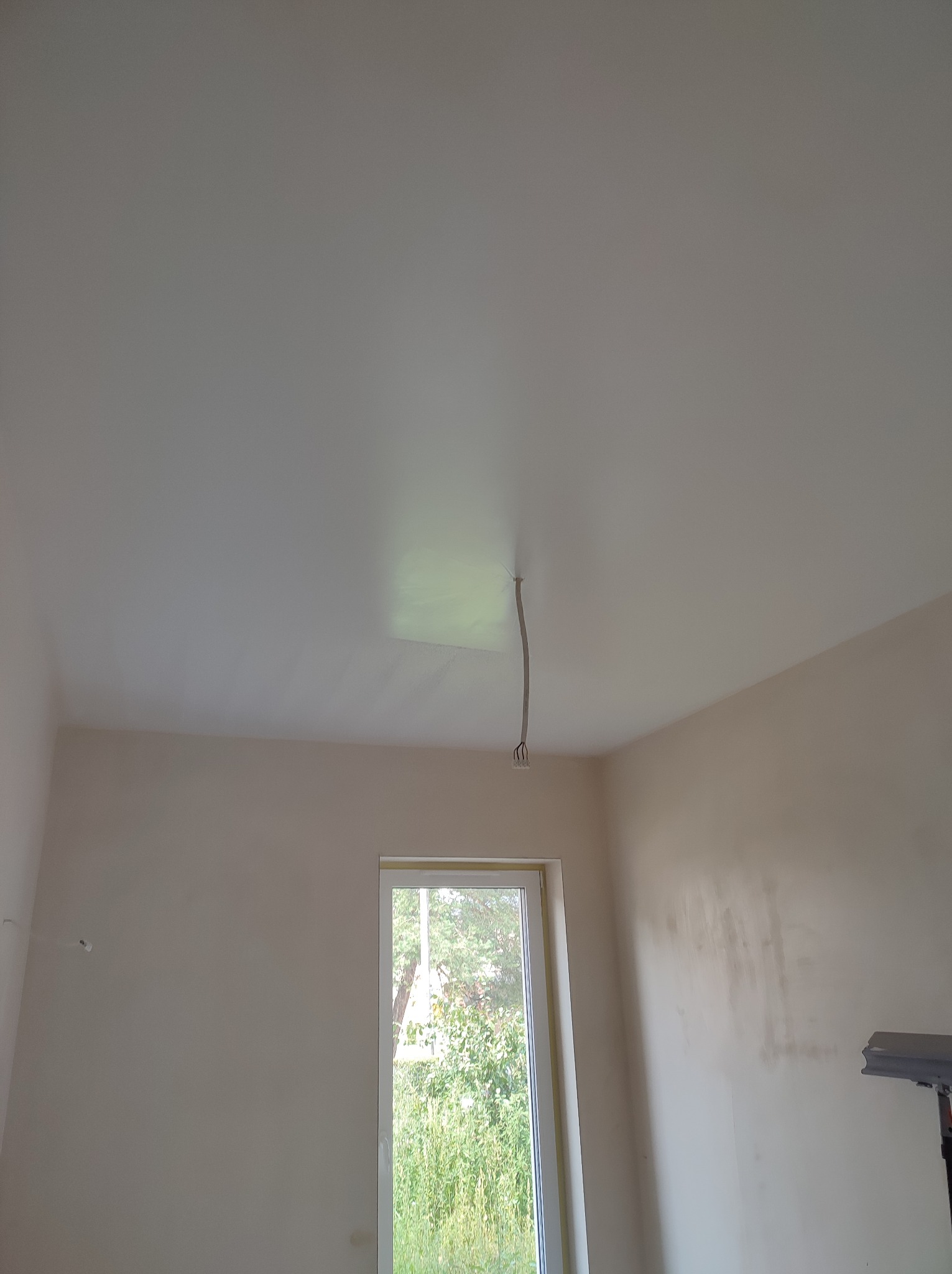 Wnętrze pokoju po szpachlowaniu, widoczne ślady po łataniu ścian, sufit przygotowany do malowania, wystający kabel elektryczny z sufitu, okno z widokiem na zieleń.