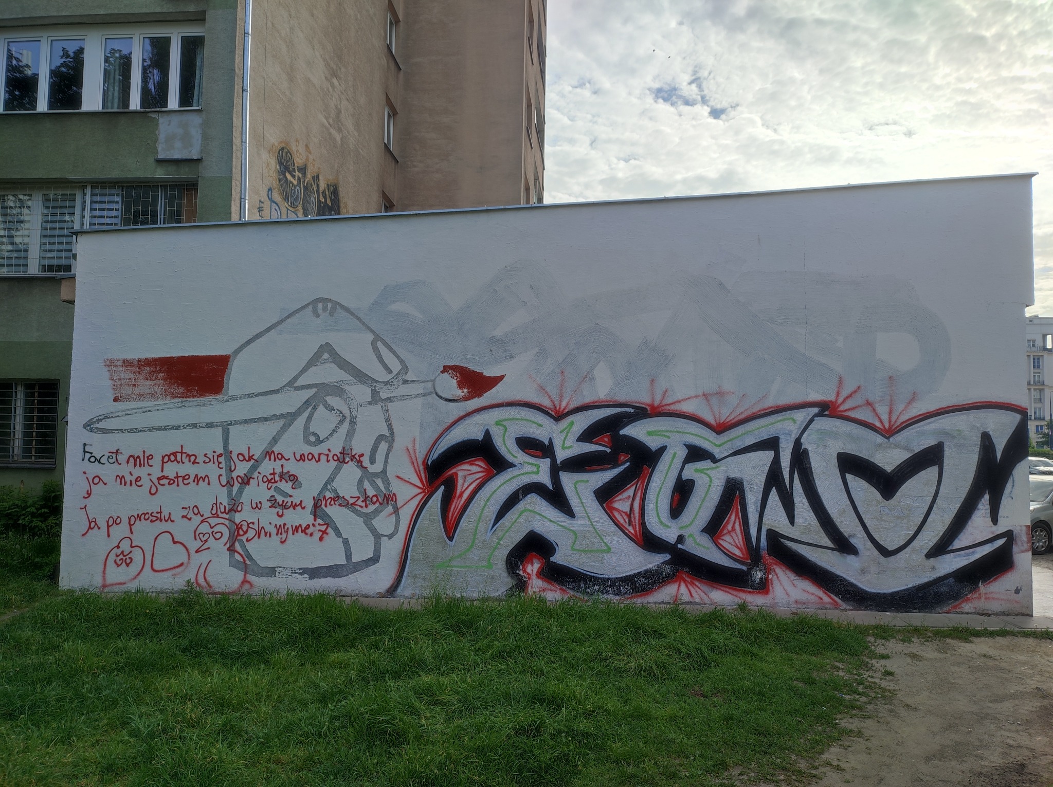 Biała ściana z graffiti przedstawiającym częściowo zamalowany szkic postaci z pędzlem oraz kolorowy napis w stylu graffiti, z tekstem po polsku napisanym czerwoną farbą, widok z poziomu trawnika.