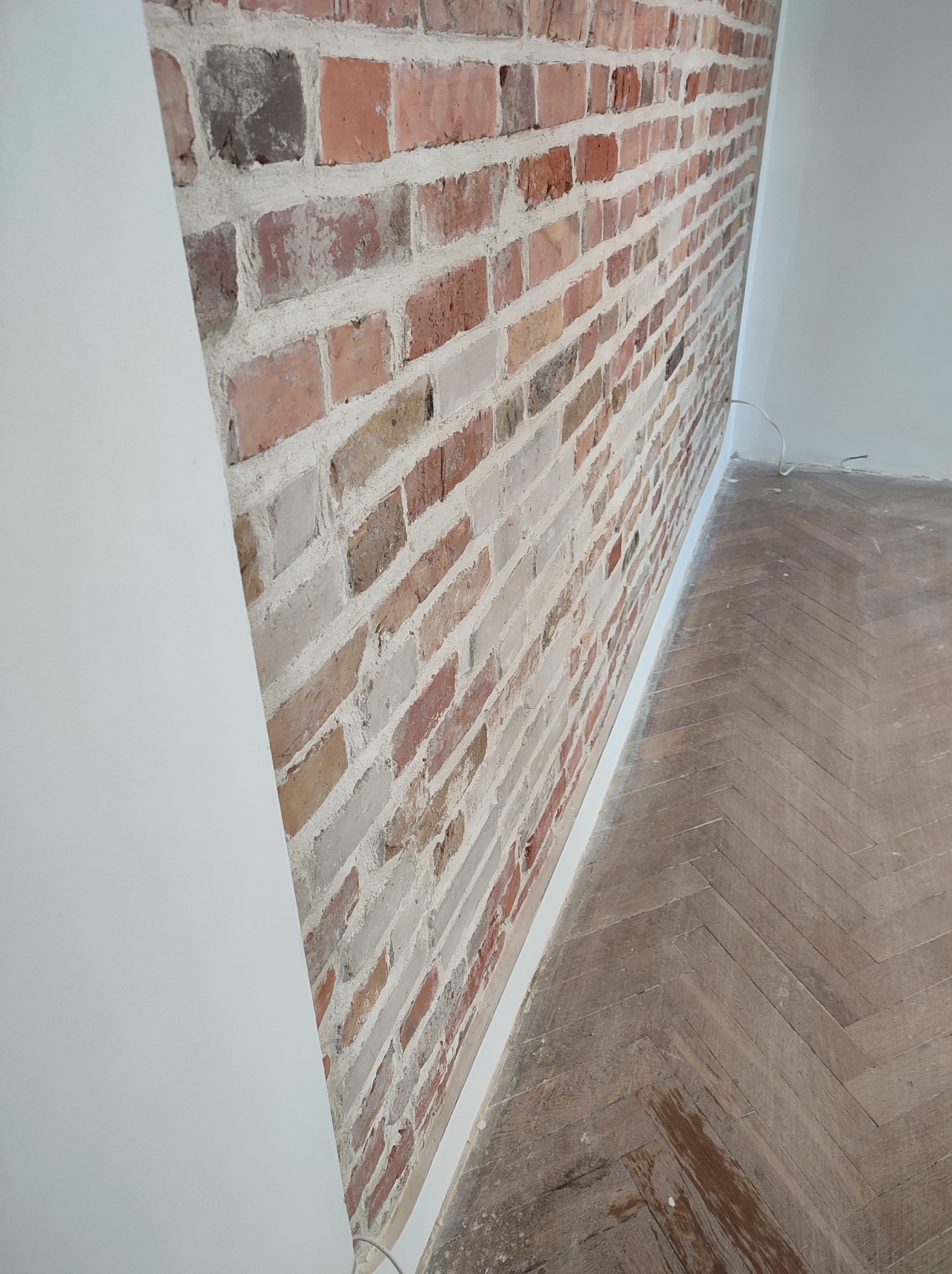 Częściowo wykończone wnętrze z ceglaną ścianą i parkietem ułożonym w jodełkę, widoczne przewody elektryczne przy podłodze.