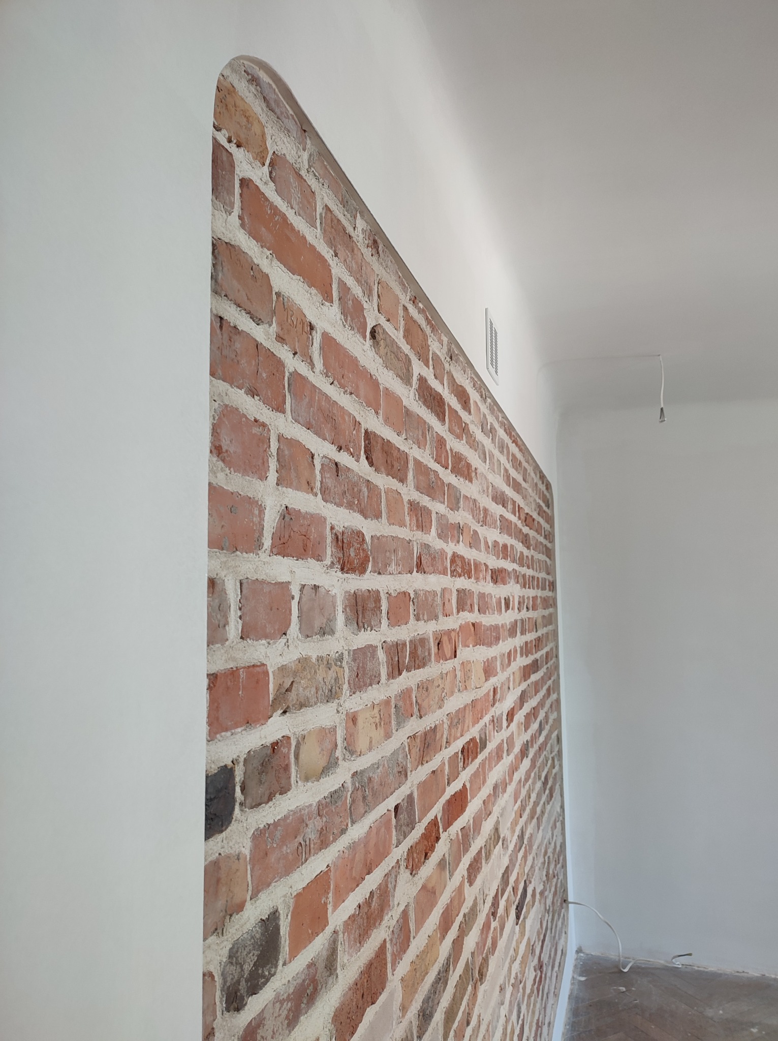 Fragment wnętrza połączonego z odnowioną ścianą z czerwonej cegły i gładko wykończoną, pomalowaną na biało ścianą z zaokrąglonym narożnikiem.