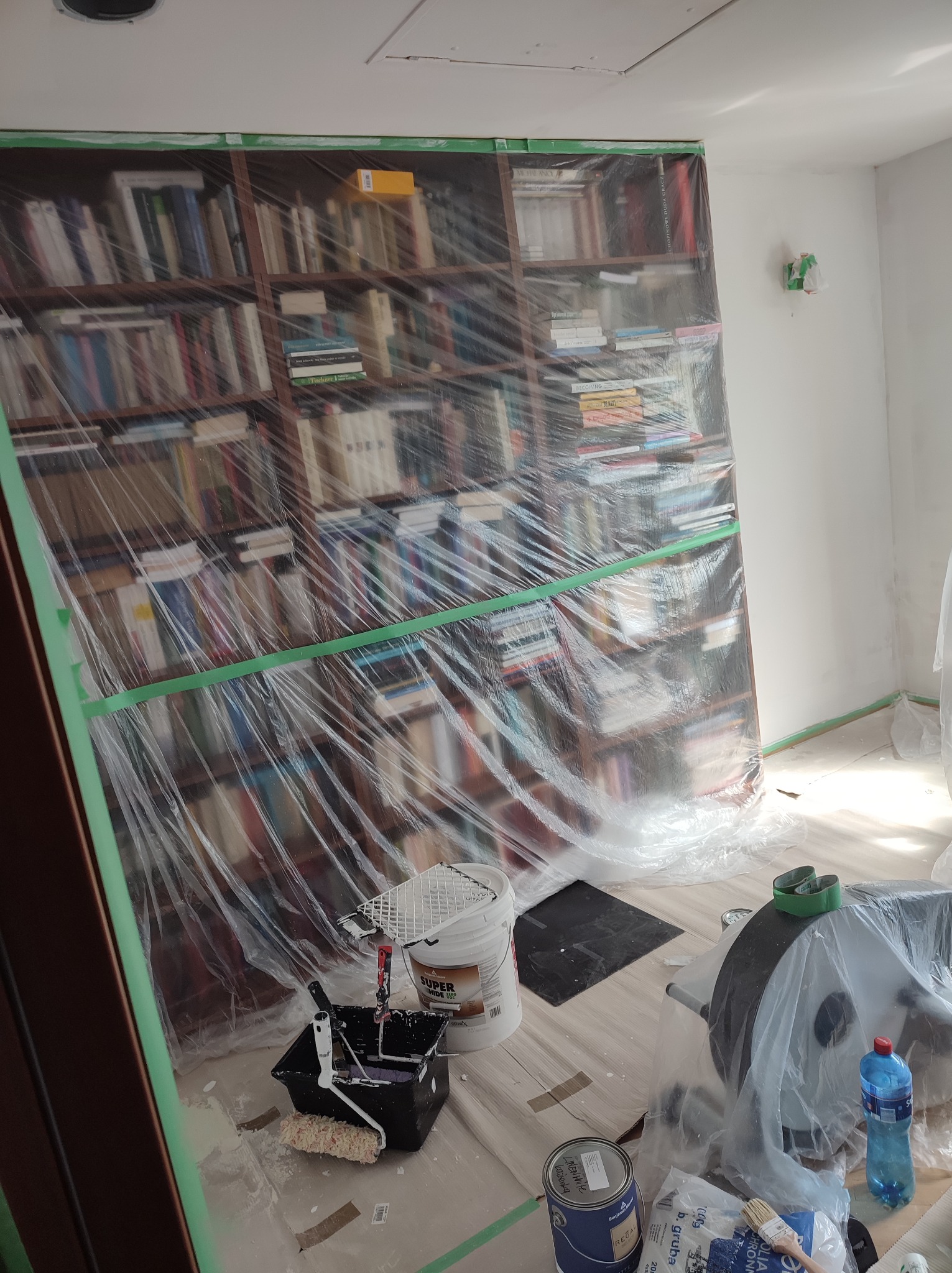 Przygotowanie do odświeżenia ścian: regał z książkami zabezpieczony folią malarską, wiadro z farbą, kuweta z wałkiem i pędzle na podłodze, zabezpieczonej folią. Widoczne taśmy malarskie przy...