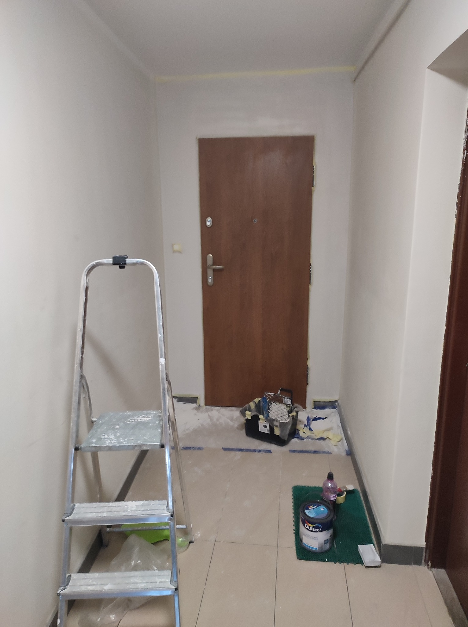 Korytarz w trakcie malowania: drabina aluminiowa, wiadro farby Dulux na zielonej wycieraczce, zabezpieczona folią podłoga, świeżo pomalowane ściany z taśmą malarską pod sufitem.