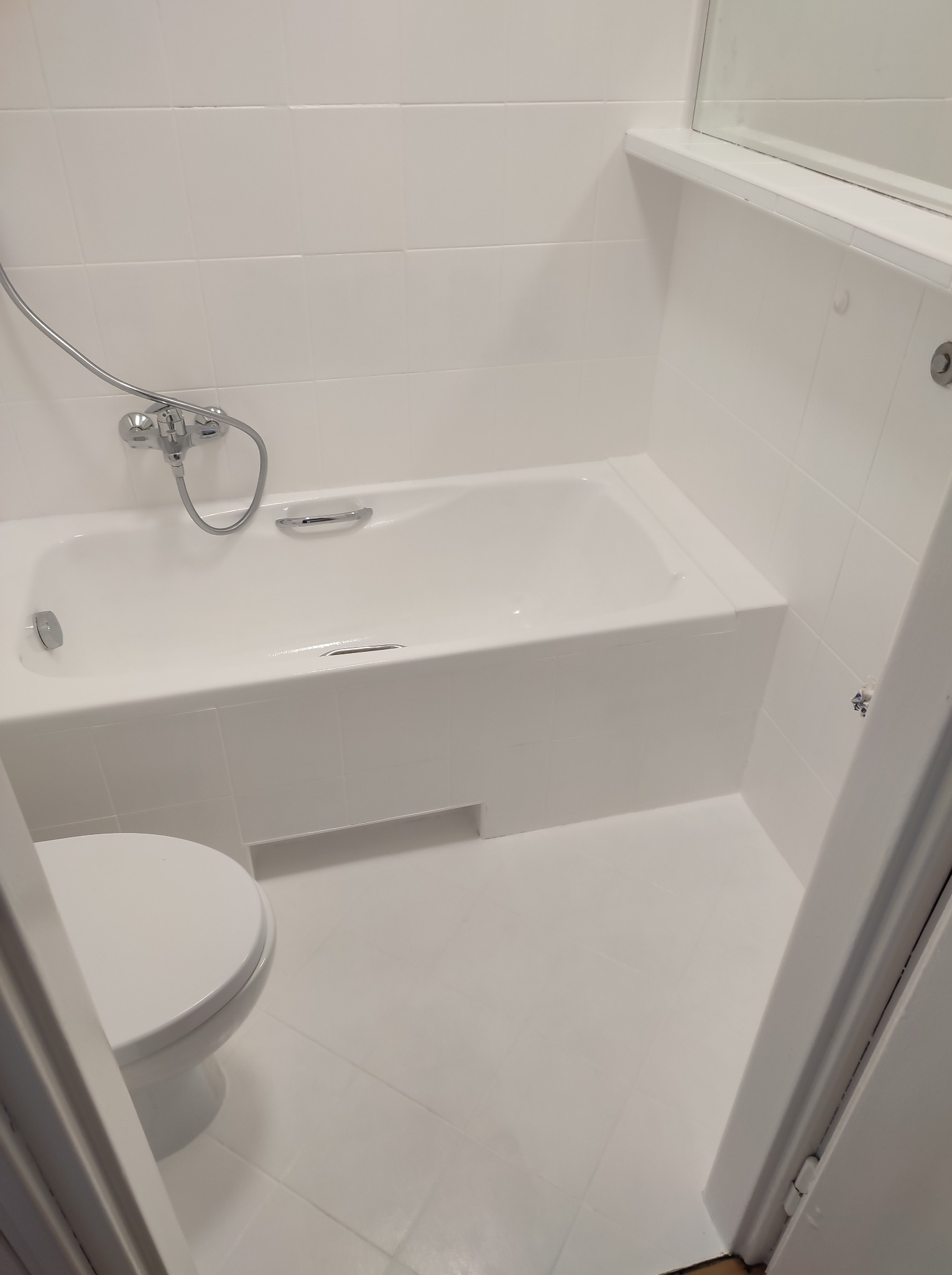 Całkowicie biała łazienka z wanną, toaletą i białymi kafelkami, świeżo po remoncie, widoczne ślady farby na podłodze.