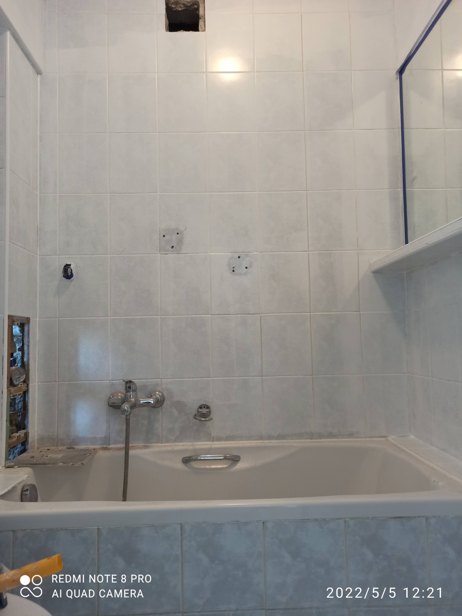 Łazienka w trakcie prac remontowych: białe płytki częściowo usunięte, odsłonięte fragmenty ściany, widoczna wanna i bateria. U góry ściany duży otwór. Widoczne ślady po wierceniach.