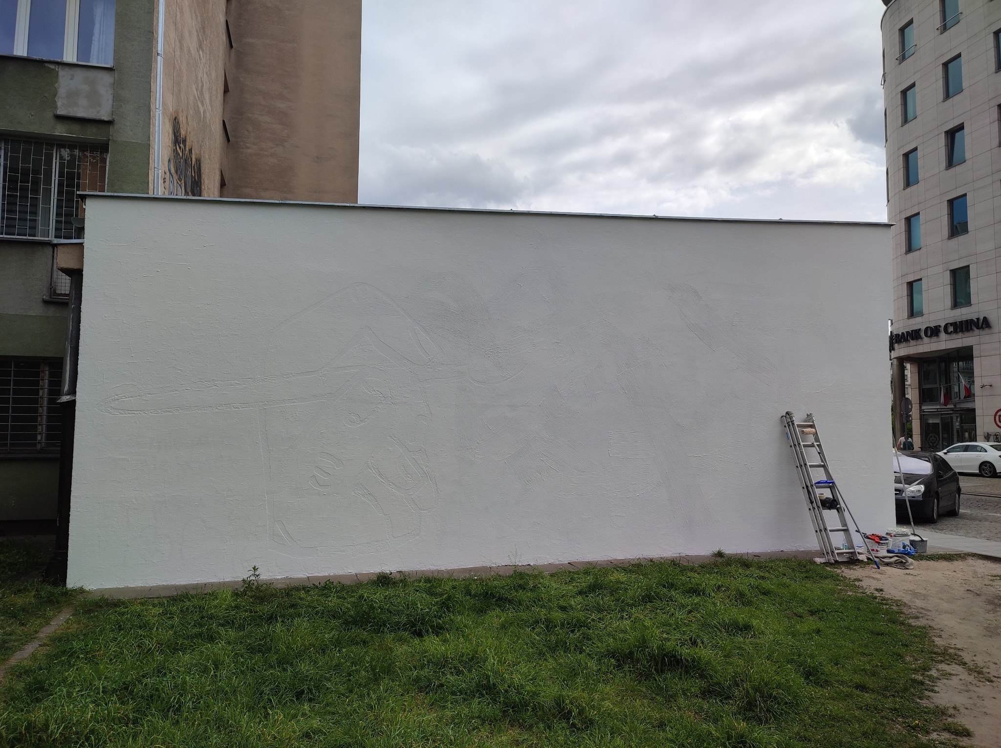 Świeżo pomalowana, biała ściana zewnętrzna budynku z widocznymi niedoskonałościami faktury, drabina oparta o ścianę, obok pojemniki z farbą i narzędzia malarskie, trawa u podstawy ściany, budynki...