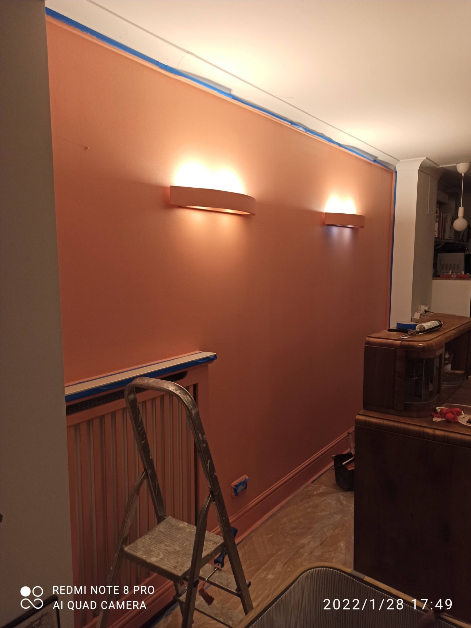 Świeżo pomalowana ściana w odcieniach łososiowego różu, zabezpieczona taśmą malarską przy suficie i listwach, z widocznymi dwoma nowoczesnymi kinkietami, drabina oparta o grzejnik, podłoga...