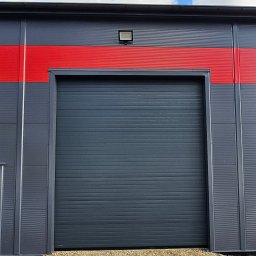 BRAMIX Szymon Wyrwa - Szara segmentowa brama garażowa z poziomymi przetłoczeniami, zamontowana w szarym budynku przemysłowym z czerwonym pasem, oświetlona zewnętrzną lampą, widok z poziomu gruntu.