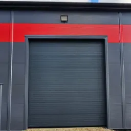 Szara segmentowa brama garażowa z poziomymi przetłoczeniami, zamontowana w szarym budynku przemysłowym z czerwonym pasem, oświetlona zewnętrzną lampą, widok z poziomu gruntu.