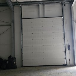 BRAMIX Szymon Wyrwa - Zamknięta segmentowa brama przemysłowa w hali produkcyjnej, widoczne panele z poziomymi przetłoczeniami, czarne uszczelki, sterownik na ścianie, surowa betonowa posadzka z zabrudzeniami.