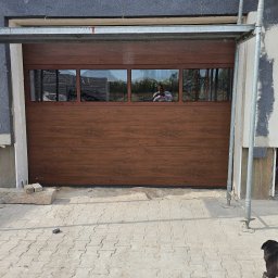 BRAMIX Szymon Wyrwa - Brązowa segmentowa brama garażowa z poziomymi panelami i czterema prostokątnymi oknami, widoczna na tle budynku w trakcie prac wykończeniowych, z rusztowaniem obok i osobą w otwartym wejściu...