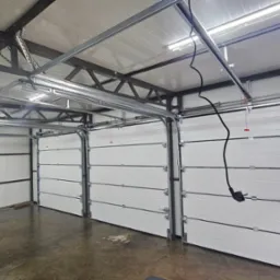 Wnętrze garażu z trzema segmentowymi bramami w kolorze białym, widoczne elementy konstrukcyjne sufitu i automatyka do bram z podłączonym kablem zasilającym.