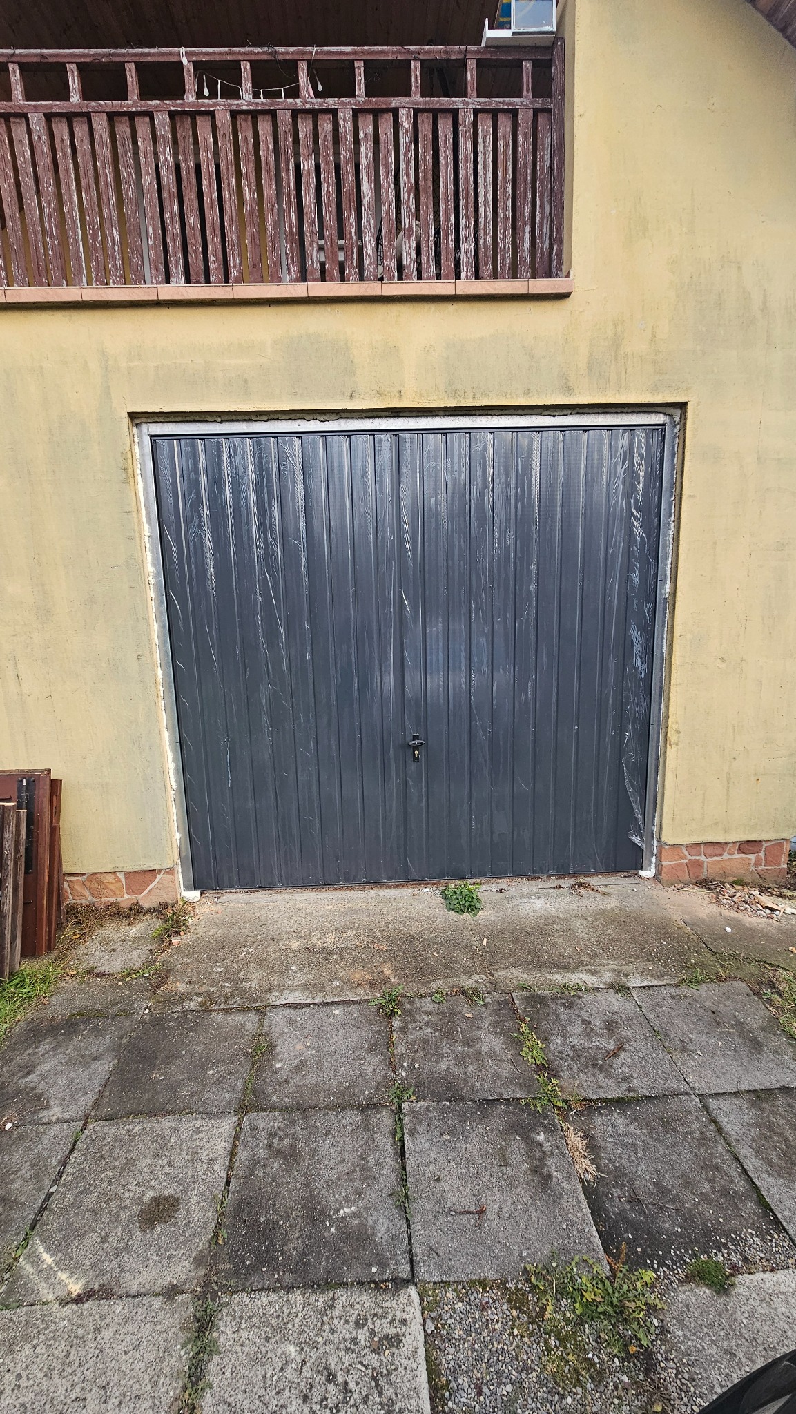 Szara brama garażowa segmentowa, zabezpieczona folią ochronną po montażu, widoczna z zewnątrz budynku z żółtą elewacją i drewnianym balkonem na piętrze.