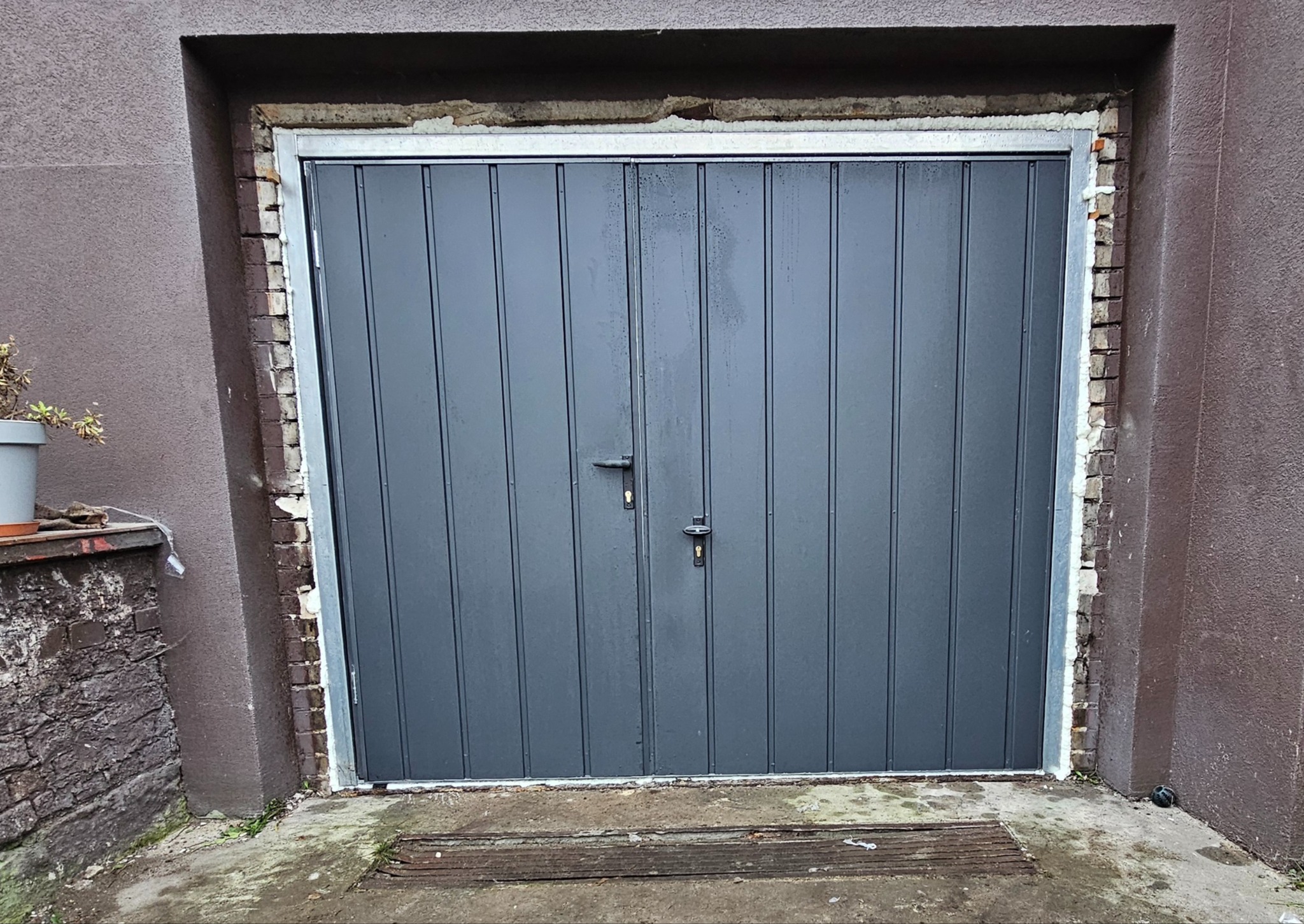 Szara, dwuskrzydłowa brama garażowa z pionowymi panelami, zamontowana w surowym, ceglano-tynkowym otworze z widoczną pianką montażową i metalową ramą, uchwycona z bliskiej perspektywy.