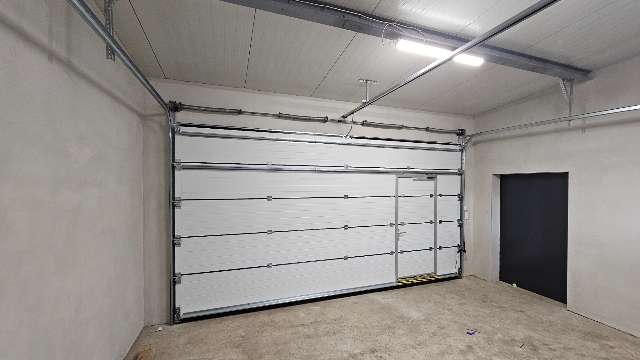 Biała segmentowa brama garażowa z drzwiami przejściowymi i żółto-czarnymi pasami ostrzegawczymi u dołu, zamontowana w surowym garażu z betonową podłogą i czarnymi drzwiami po prawej stronie.