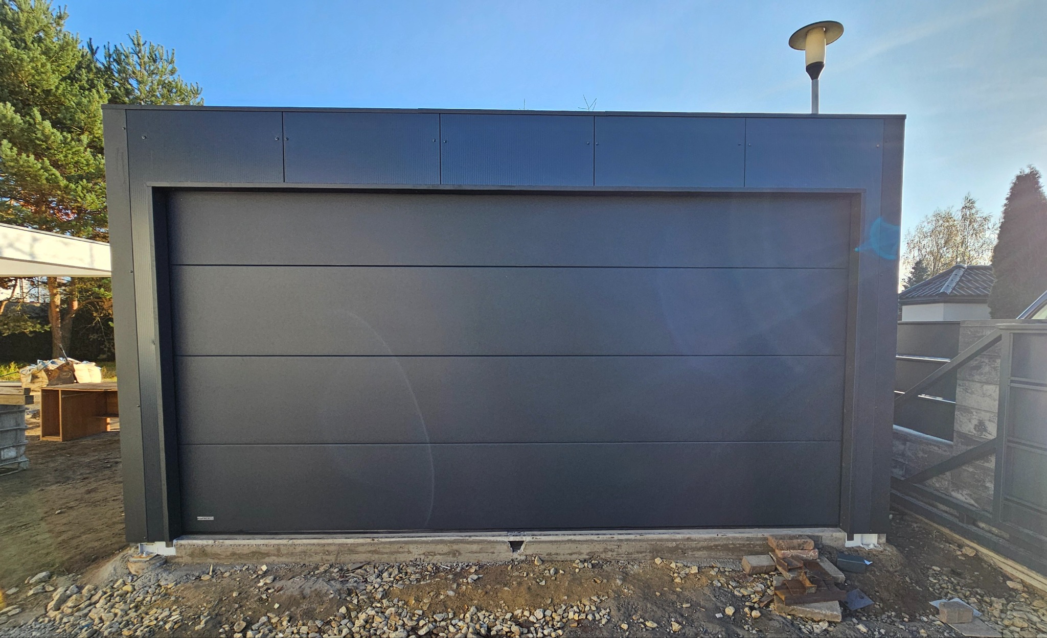 Nowoczesna, segmentowa, grafitowa brama garażowa z poziomymi przetłoczeniami, zamontowana w ciemnej obudowie z oświetleniem zewnętrznym, widok z zewnątrz na tle nieba i zieleni.