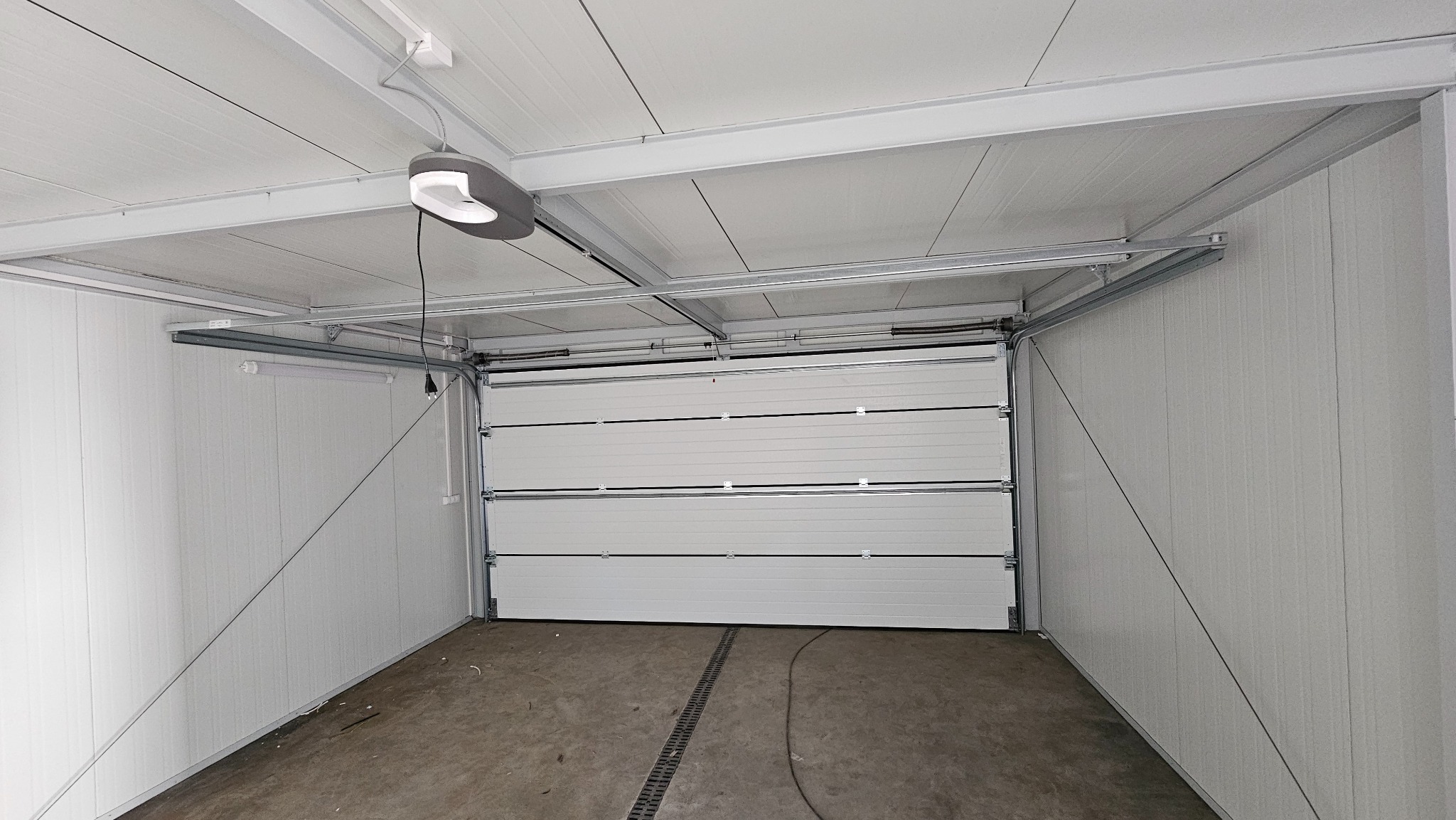 Wnętrze garażu z częściowo otwartą białą segmentową bramą, widoczny napęd sufitowy z oświetleniem LED i szyny prowadzące bramę.