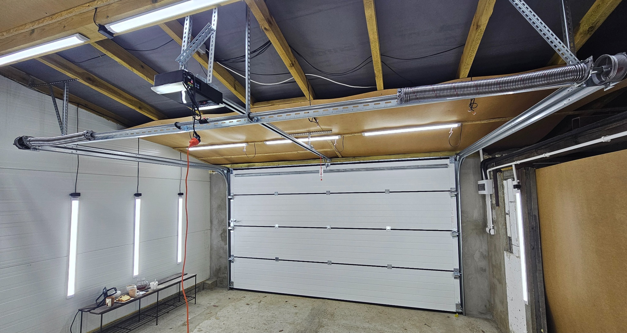 Wnętrze garażu z białą segmentową bramą, widoczne oświetlenie sufitowe, mechanizm sprężynowy bramy i automatyka. Na ścianie wiszą lampy, a pod nimi stoi stolik z kawą i przekąskami.