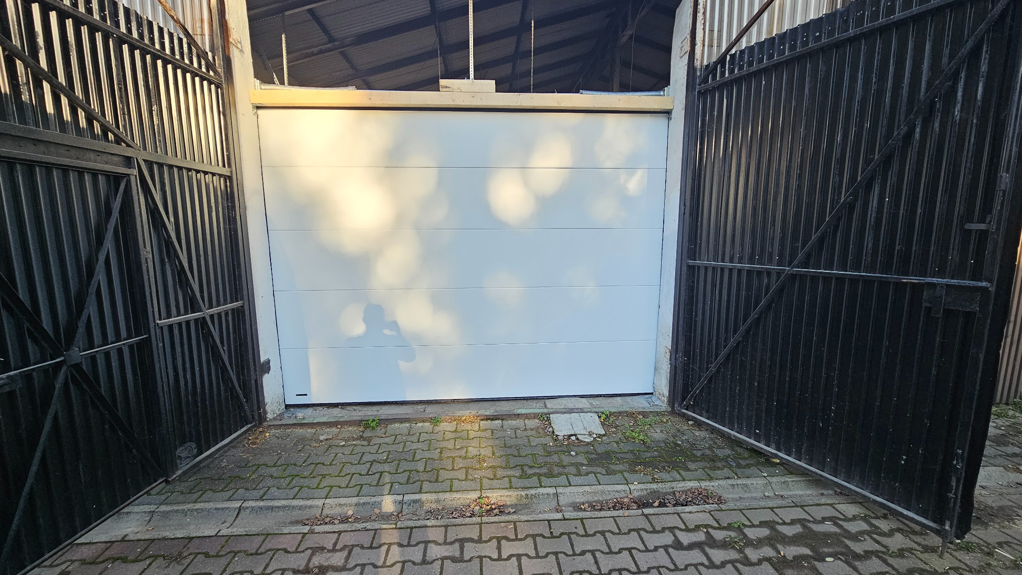 Otwarta, biała, segmentowa konstrukcja z widocznym mechanizmem na górze, umieszczona w ciemnym garażu z otwartymi, metalowymi, czarnymi wrotami po bokach i kostką brukową na podjeździe.