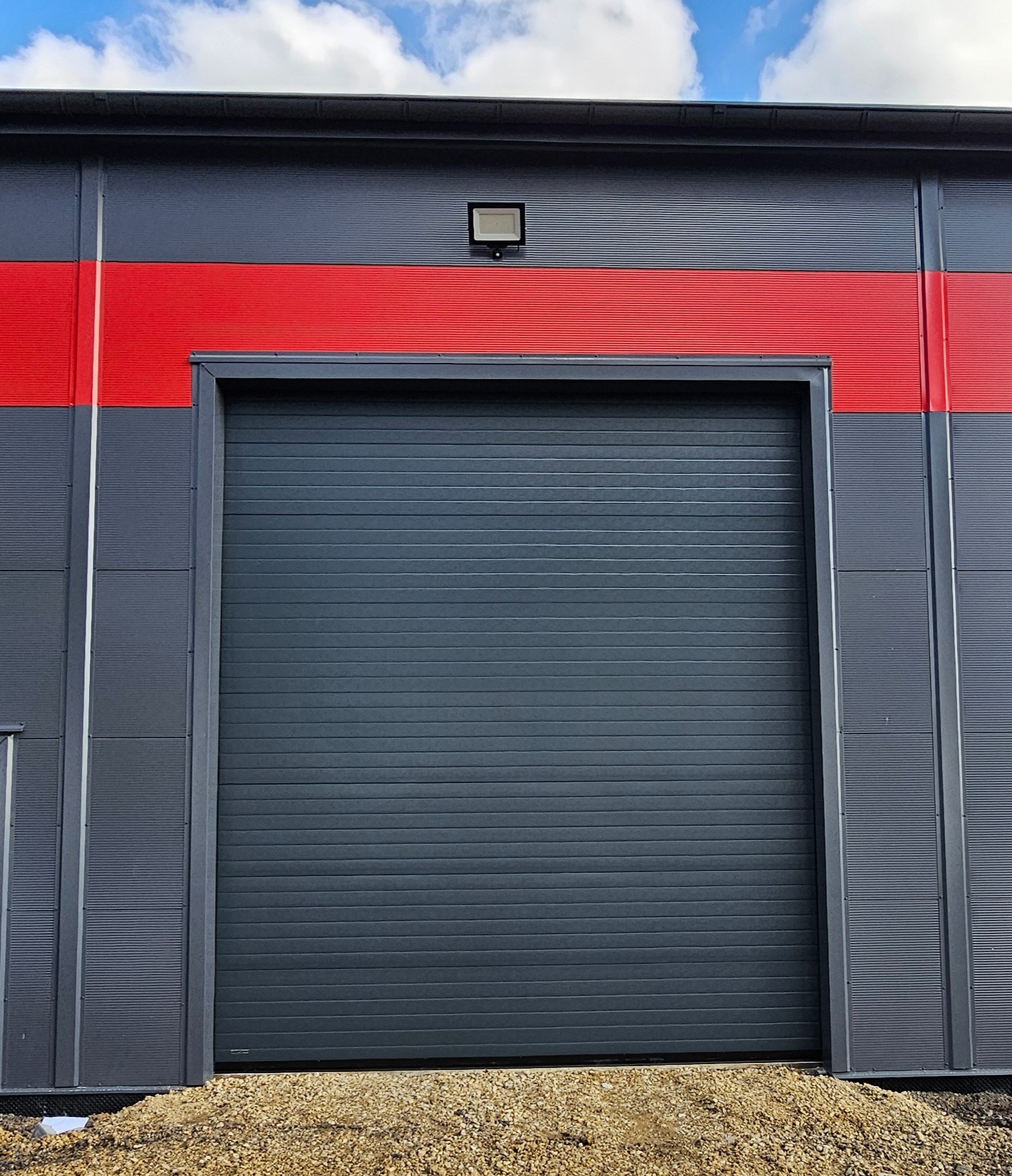 Szara segmentowa brama garażowa z poziomymi przetłoczeniami, zamontowana w szarym budynku przemysłowym z czerwonym pasem, oświetlona zewnętrzną lampą, widok z poziomu gruntu.