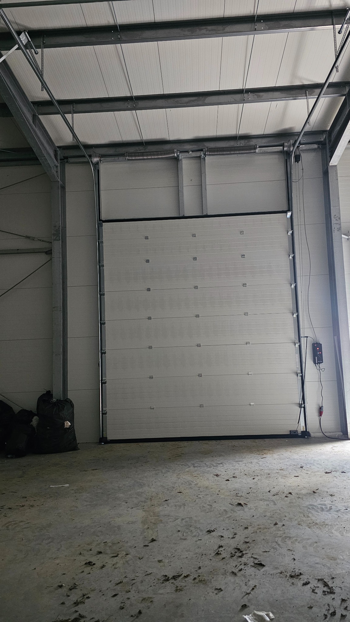 Zamknięta segmentowa brama przemysłowa w hali produkcyjnej, widoczne panele z poziomymi przetłoczeniami, czarne uszczelki, sterownik na ścianie, surowa betonowa posadzka z zabrudzeniami.