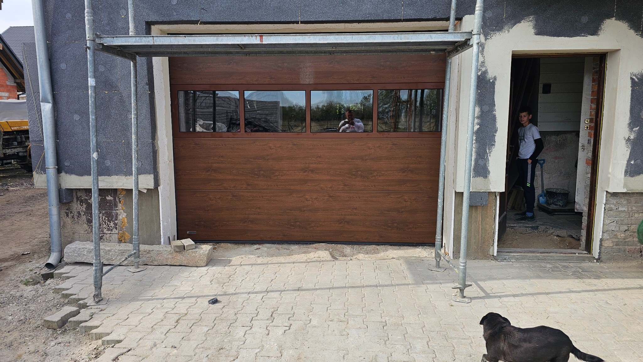 Brązowa segmentowa brama garażowa z poziomymi panelami i czterema prostokątnymi oknami, widoczna na tle budynku w trakcie prac wykończeniowych, z rusztowaniem obok i osobą w otwartym wejściu...