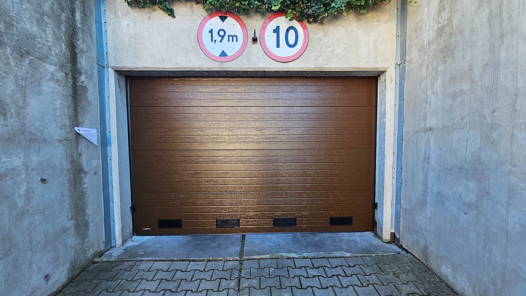 Zamknięta, brązowa brama segmentowa w Koniecpolu, z widocznymi znakami ograniczenia wysokości 1,9m i prędkości 10km/h nad nią, oraz fragmentem bluszczu.