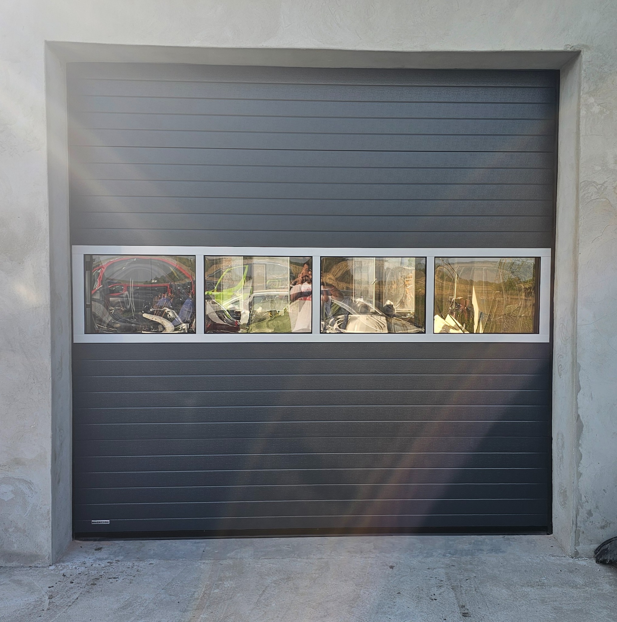 Szara segmentowa brama garażowa z czterema prostokątnymi okienkami, przez które widać zarysowane kształty przedmiotów i roślin wewnątrz garażu.