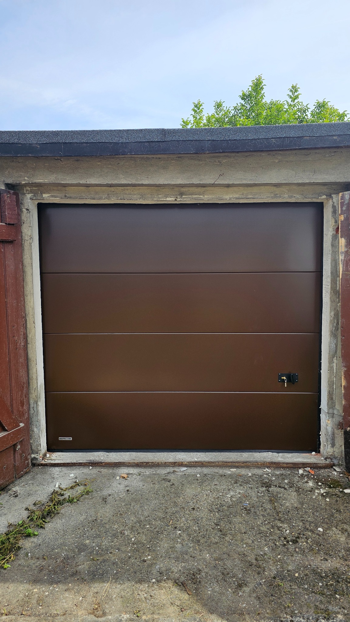 Ciemnobrązowa brama garażowa segmentowa, z małym zamkiem na klucz, widziana od zewnątrz, z widocznymi elementami betonowej konstrukcji garażu i fragmentem roślinności na górze.