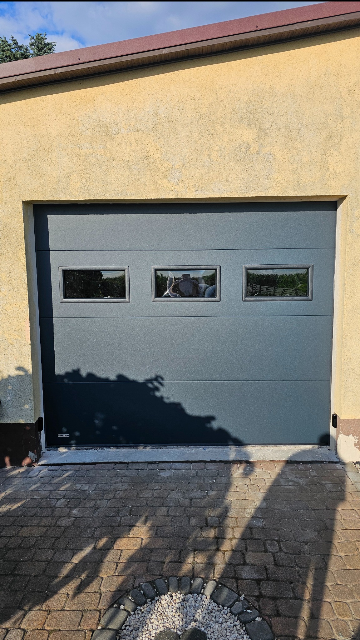 Szara segmentowa brama garażowa z trzema małymi okienkami, widoczna z zewnątrz na tle żółtej ściany budynku, cień drzewa na bramie i kostce brukowej.