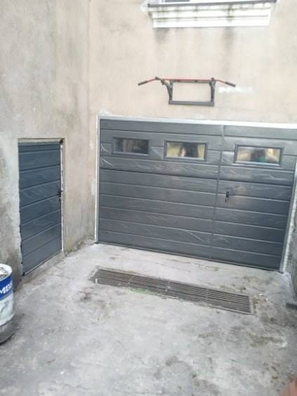 Szara segmentowa brama garażowa z oknami, zamontowana obok wąskich drzwi wejściowych, widoczne rury pod sufitem i kratka ściekowa w posadzce.