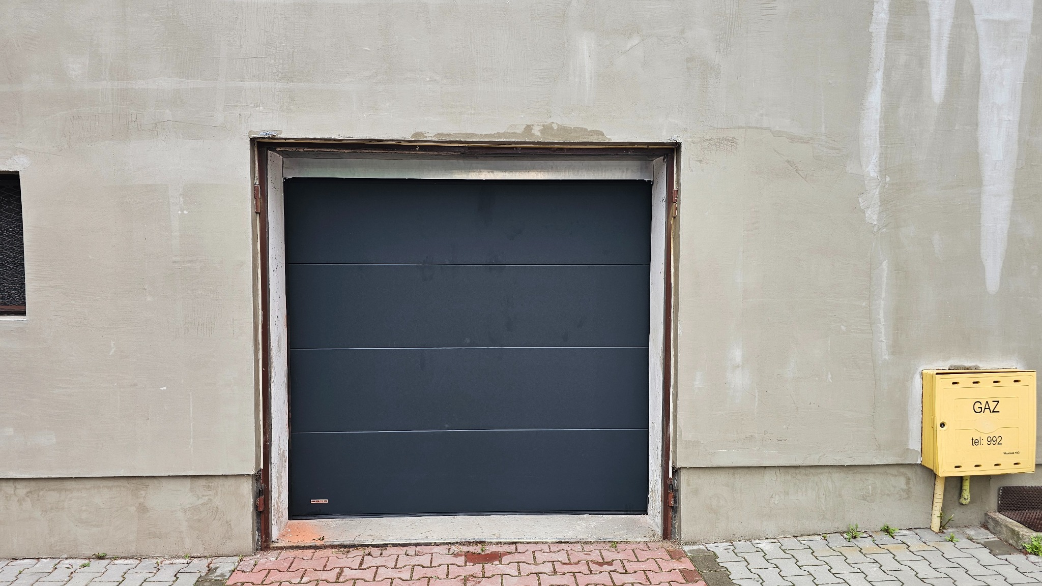 Zamknięta, segmentowa brama garażowa w kolorze grafitowym, widoczna z zewnątrz, z żółtą skrzynką gazową po prawej stronie na tle szarego, surowego muru.