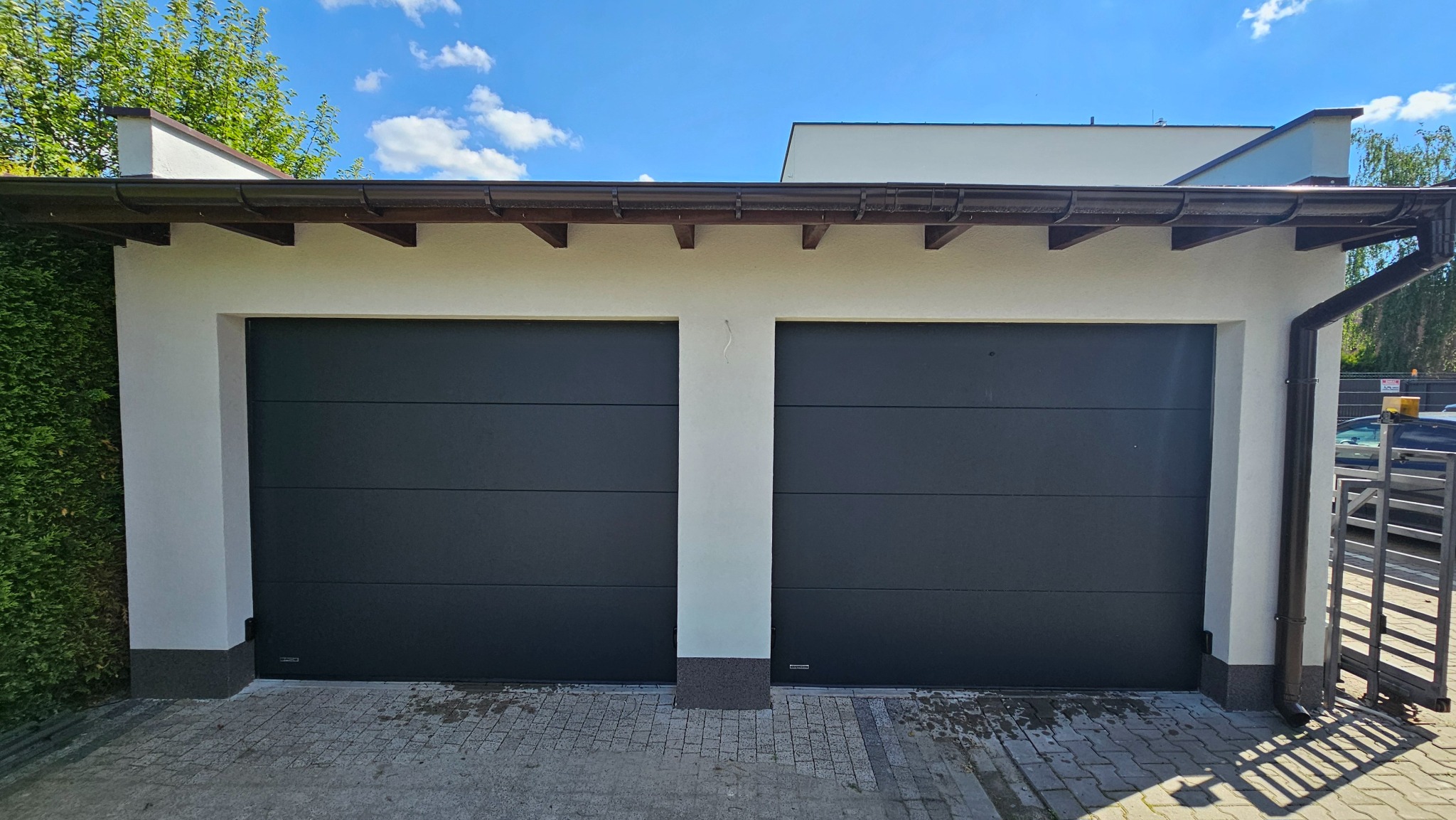 Dwustanowiskowy garaż z czarnymi segmentowymi drzwiami, białą elewacją i kostką brukową przed wjazdem, widok z zewnątrz w słoneczny dzień.