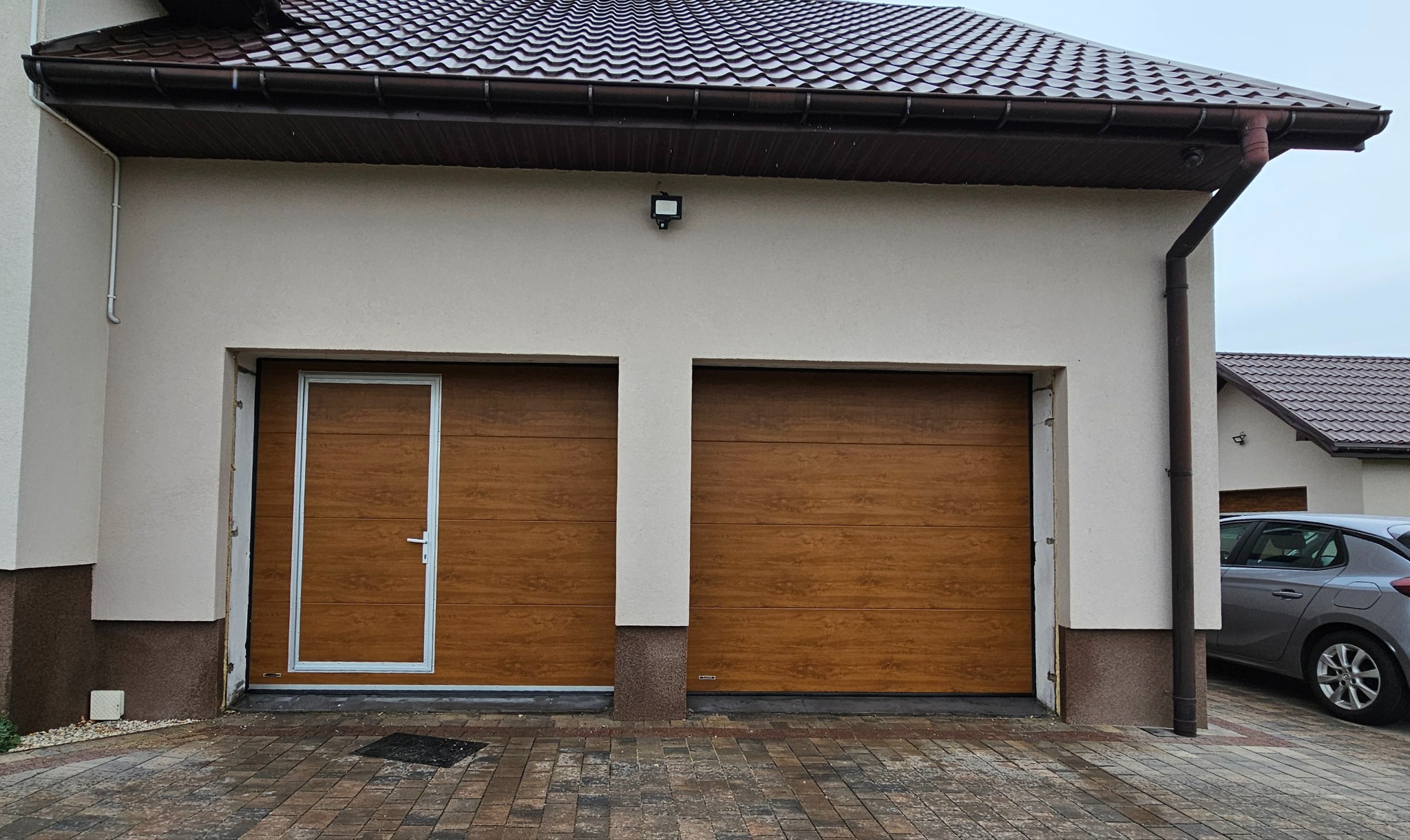 Dwustanowiskowy garaż z brązowymi segmentowymi drzwiami, zintegrowanymi drzwiami wejściowymi po lewej stronie, na zewnątrz domu z brązowym dachem i kostką brukową.