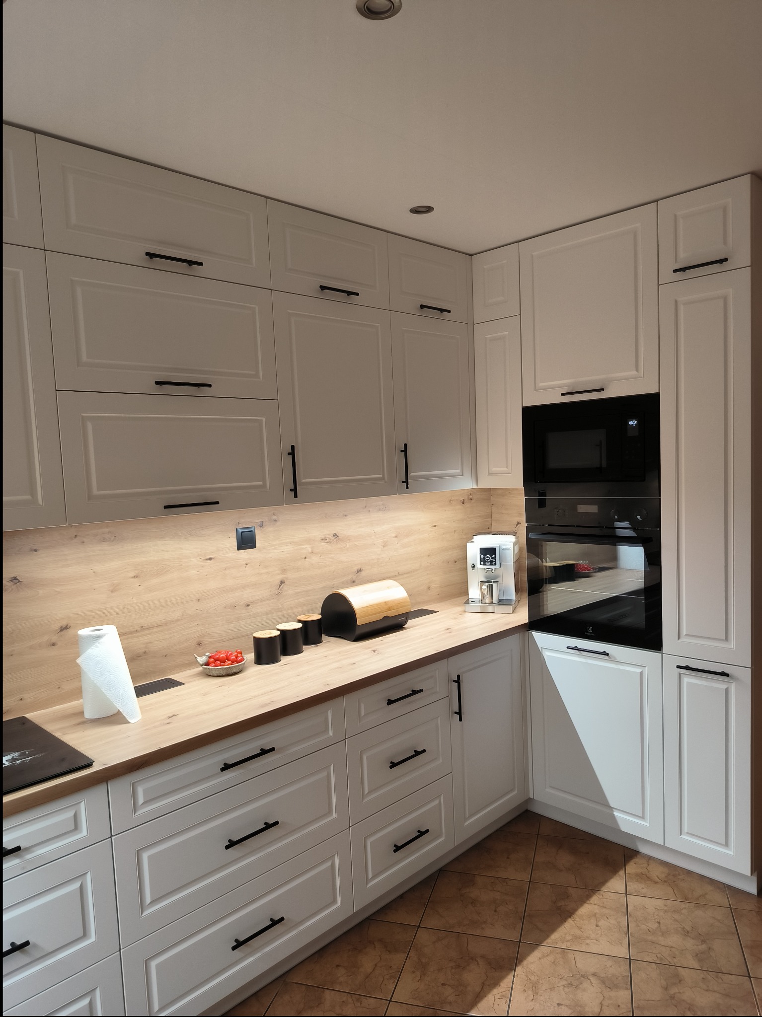 Jasna kuchnia z frezowanymi frontami, czarnymi uchwytami, drewnianym blatem i sprzętem AGD w zabudowie. Na blacie ekspres do kawy, pojemniki i miseczka z pomidorami koktajlowymi.