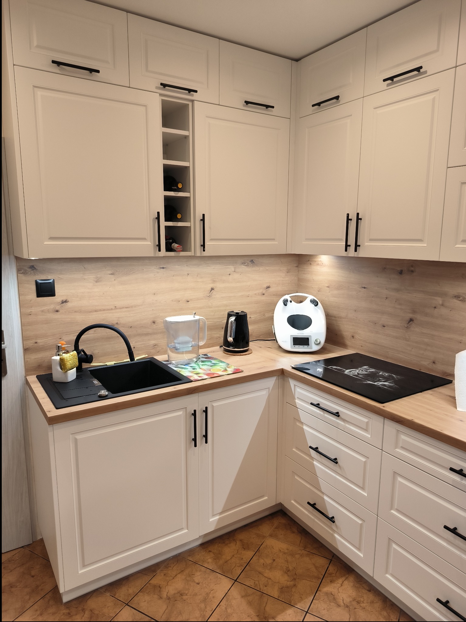 Jasna, narożna zabudowa kuchenna z frezowanymi frontami, czarnymi uchwytami i drewnianym blatem; widoczny zlew z czarną armaturą, płyta indukcyjna z grafiką, ekspres do kawy i półka na wino.