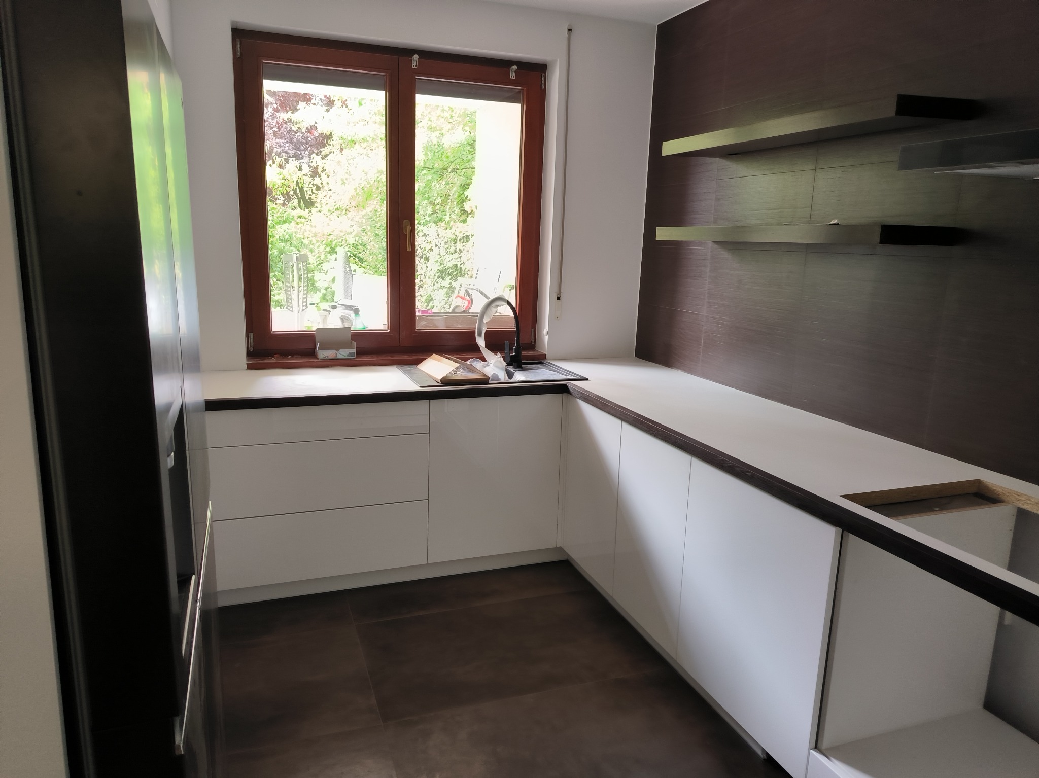 Nowoczesna, biała zabudowa kuchenna z ciemnym blatem, okno z widokiem na zieleń, brązowe płytki na ścianie i drewniane półki, etap montażu widoczny pod blatem.