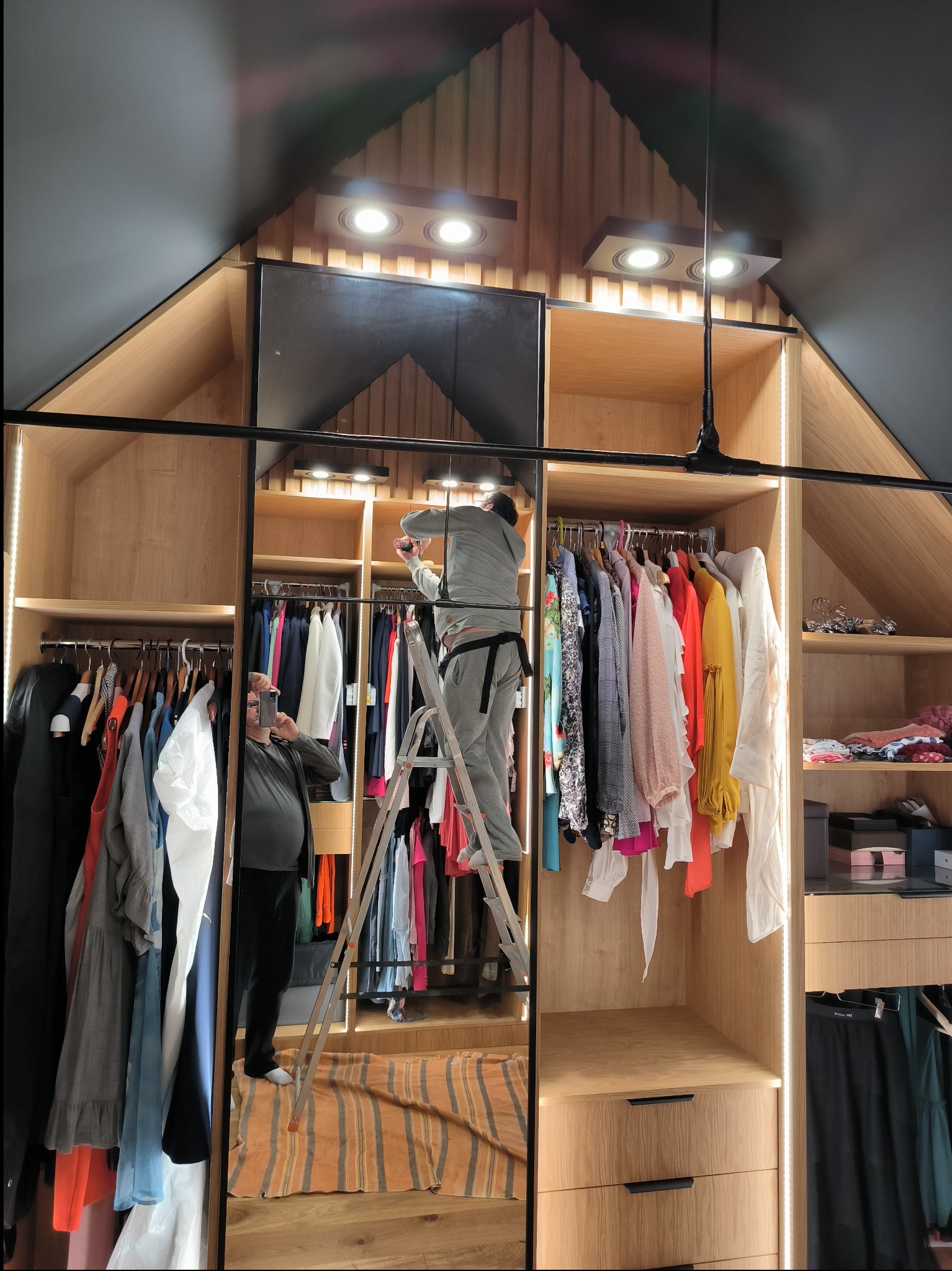 Montaż oświetlenia w szafie wnękowej na poddaszu z garderobą, widoczny mężczyzna na drabinie i odbicie fotografa.