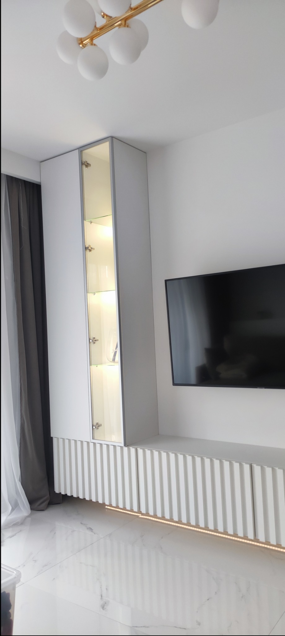 Nowoczesna, biała zabudowa meblowa z szafką z przeszklonymi drzwiami, podświetlonymi półkami i dekoracyjnym panelem ryflowanym u dołu, uzupełniona o podświetlenie LED pod szafką RTV i marmurową...