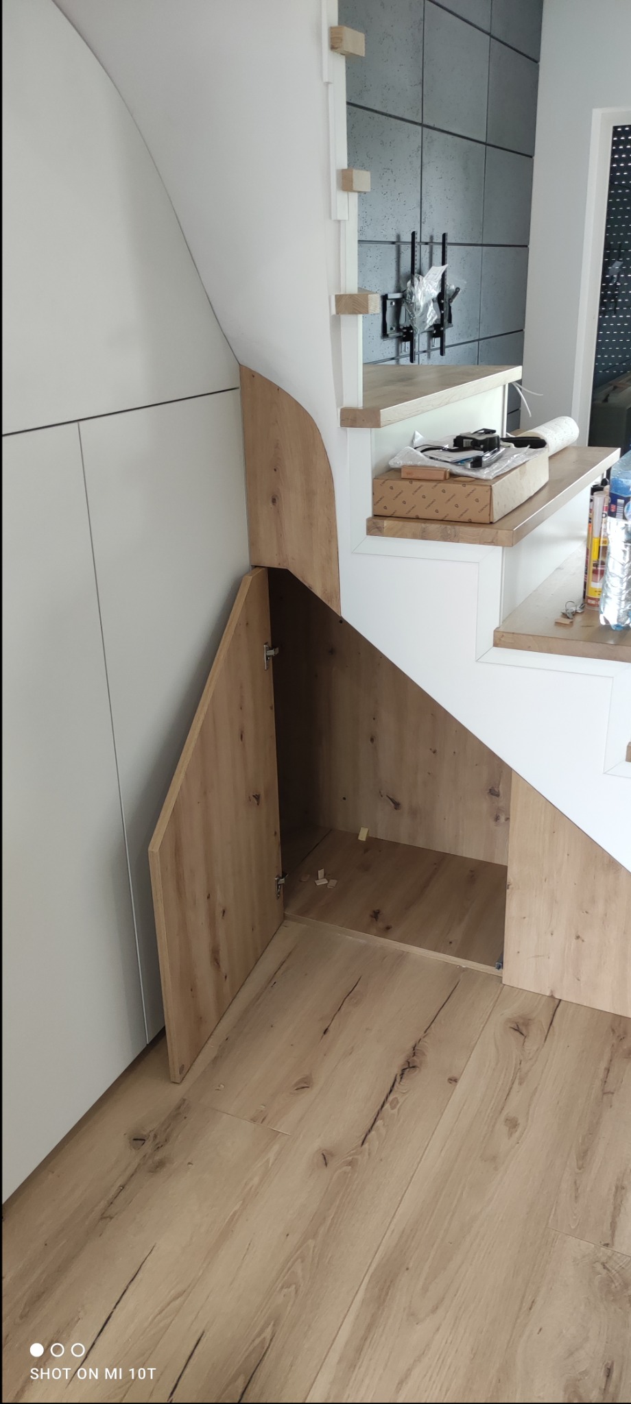 Szafka z drewnianymi drzwiczkami pod schodami, z otwartymi drzwiami, ukazująca wnętrze. Na schodach leżą narzędzia i akcesoria montażowe.