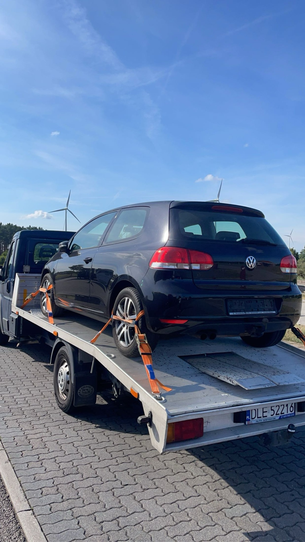 Czarny Volkswagen Golf zabezpieczony pomarańczowymi pasami na platformie lawety, w tle turbiny wiatrowe i błękitne niebo.