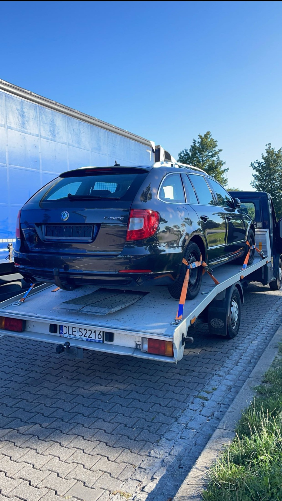 Czarny samochód Skoda Superb kombi, zabezpieczony pomarańczowymi pasami transportowymi, przewożony na platformie lawety na tle błękitnego nieba i częściowo widocznej naczepy tira.