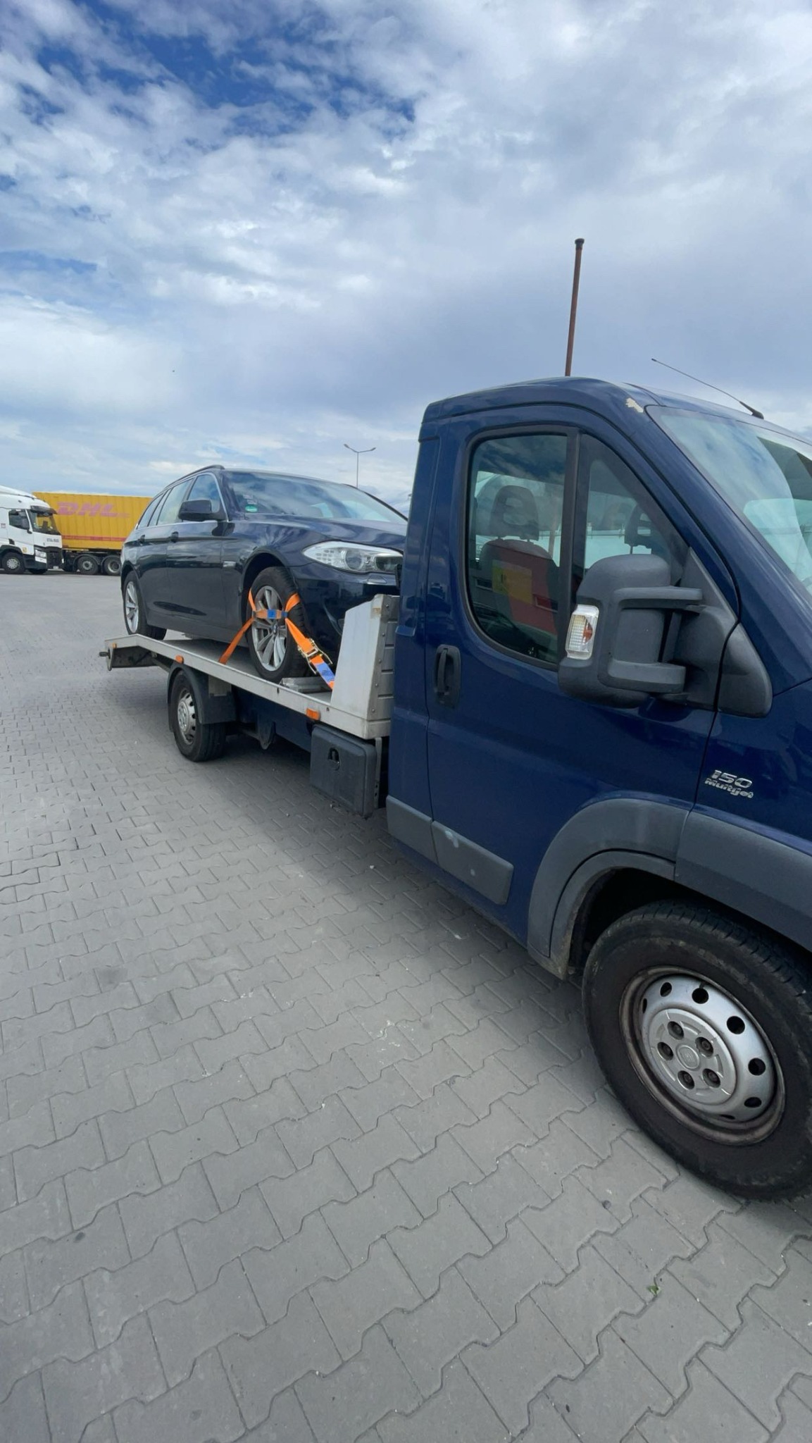 Czarny samochód osobowy zabezpieczony pomarańczowymi pasami transportowymi na platformie niebieskiej autolawety, widok z boku na tle pochmurnego nieba.