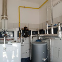 Termo Partner - Biały piec grzewczy Vaillant z szarym zasobnikiem wody, rurami w izolacji i żółtą instalacją gazową w pomieszczeniu wyłożonym częściowo kafelkami.