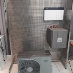 Szara pompa ciepła Nibe AMS10, składająca się z jednostki zewnętrznej z wentylatorem, modułu wewnętrznego z rurami i zasobnika, umieszczona na tle perforowanej metalowej ścianki ekspozycyjnej.