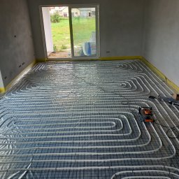 Termo Partner - Rozłożone rury ogrzewania podłogowego na siatce w surowym wnętrzu z widokiem na zewnątrz przez okno balkonowe, widoczny rozwijak do rur.