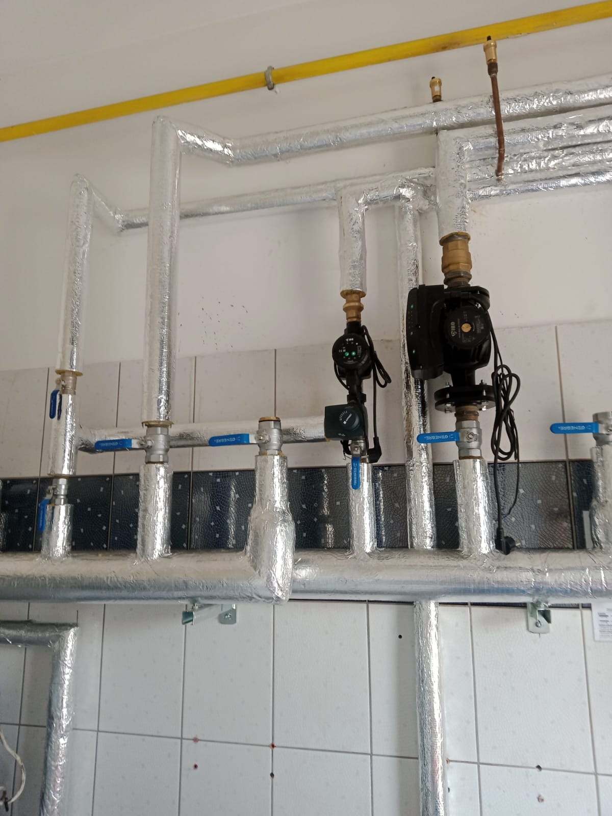 Izolowane rury grzewcze z zaworami i pompami obiegowymi zamontowane na białej ścianie z kwadratowymi płytkami, widoczna żółta rura gazowa pod sufitem.