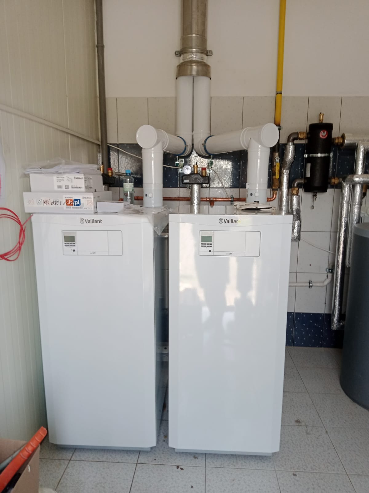 Dwa białe, wiszące piece grzewcze marki Vaillant z podłączonymi rurami, zamontowane w pomieszczeniu z wykafelkowanymi ścianami i podłogą. Na jednym z pieców leżą oryginalne opakowania.