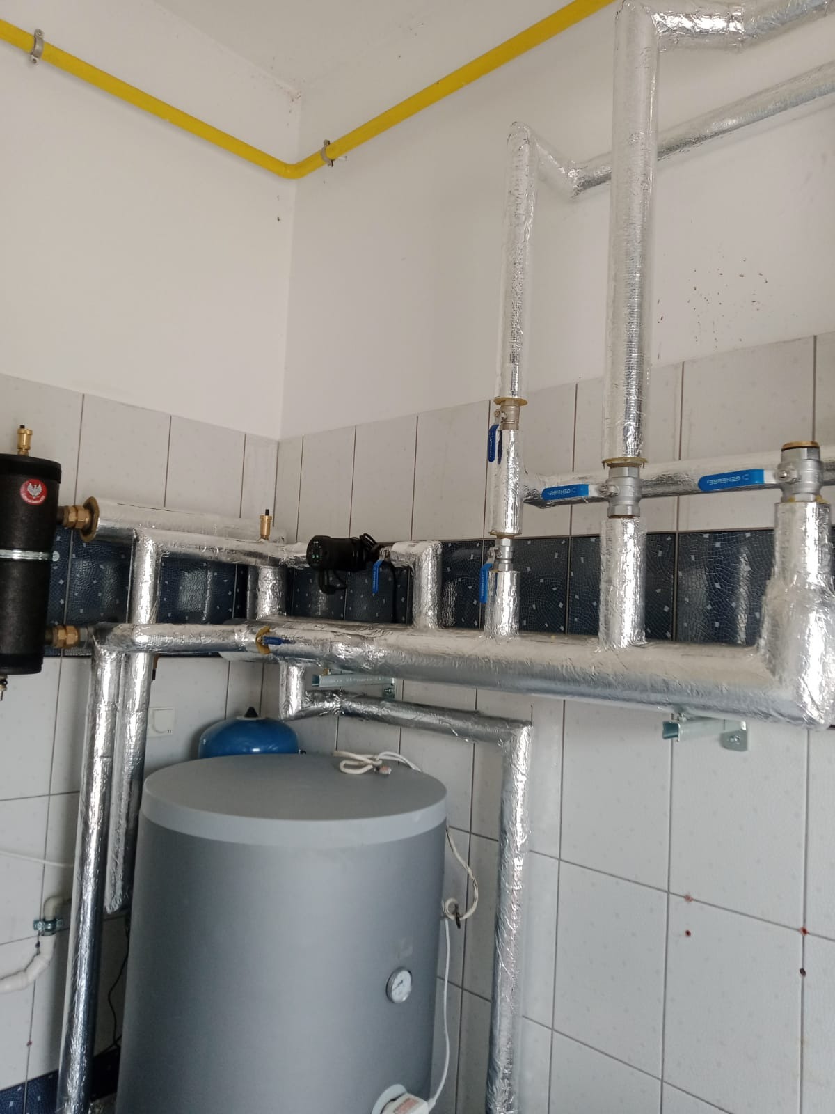 Szary zasobnik ciepłej wody użytkowej z niebieskim naczyniem wzbiorczym, połączony z instalacją grzewczą z izolowanymi rurami, zamontowany w pomieszczeniu z białymi kafelkami, widoczny żółty...
