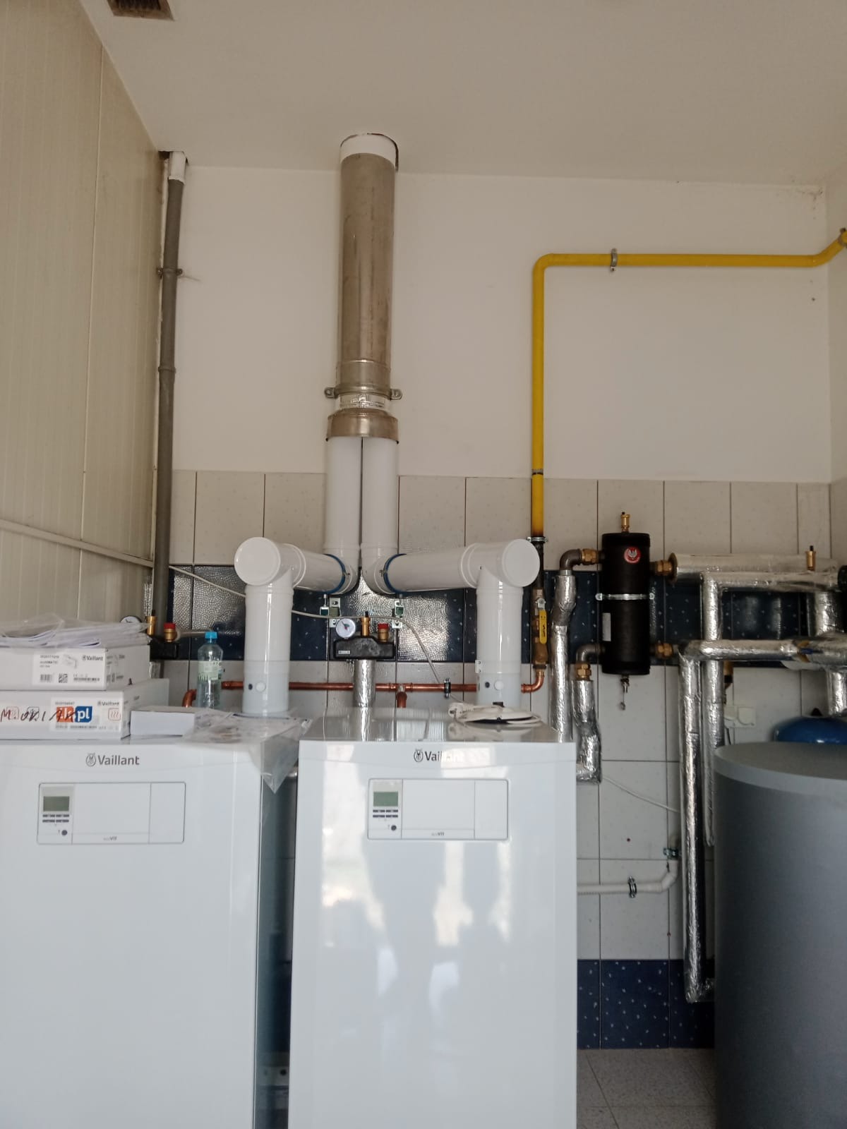 Dwa białe kotły grzewcze Vaillant z podłączonymi rurami, widoczna żółta instalacja gazowa, butelka wody i pudełka na kotłach, tło stanowią kafelki w kolorze białym i granatowym